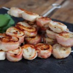 Brochetas de pechuga de pollo envueltas en beicon, doradas a la parrilla y servidas sobre tabla de pizarra con hierbas frescas.