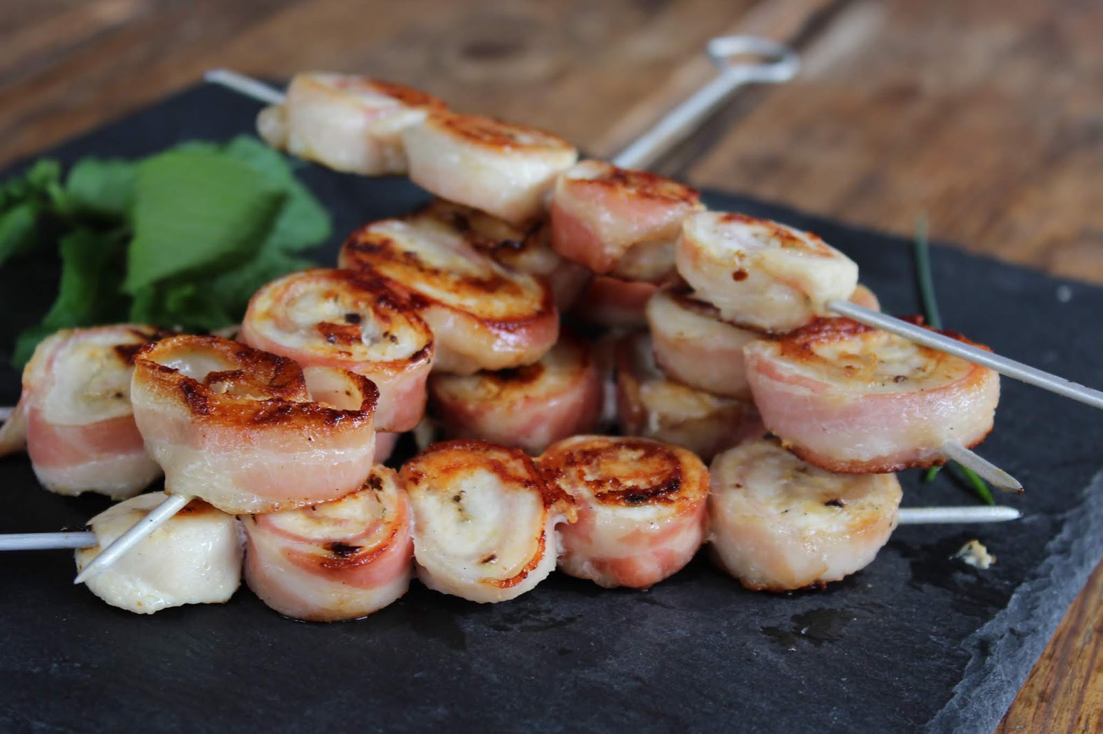 Brochetas de pechuga de pollo envueltas en beicon, doradas a la parrilla y servidas sobre tabla de pizarra con hierbas frescas.