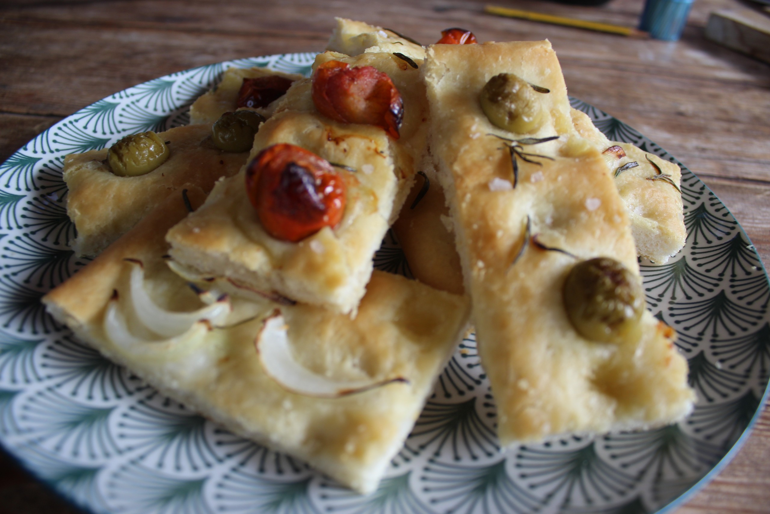 Focaccia casera italiana servida en un plato decorativo, con tomates cherry, aceitunas verdes, cebolla y romero sobre una corteza dorada y crujiente.
