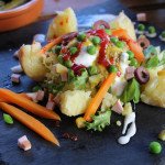 Patatas rellenas de feria servidas sobre pizarra negra, decoradas con guisantes, zanahorias, jamón, aceitunas, salsas y hojas verdes.