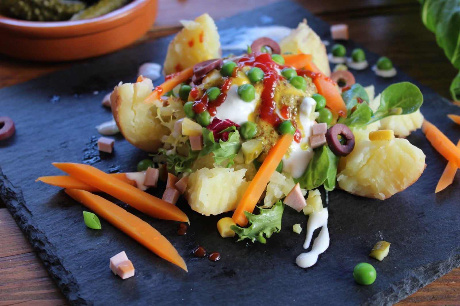 Patatas rellenas de feria servidas sobre pizarra negra, decoradas con guisantes, zanahorias, jamón, aceitunas, salsas y hojas verdes.