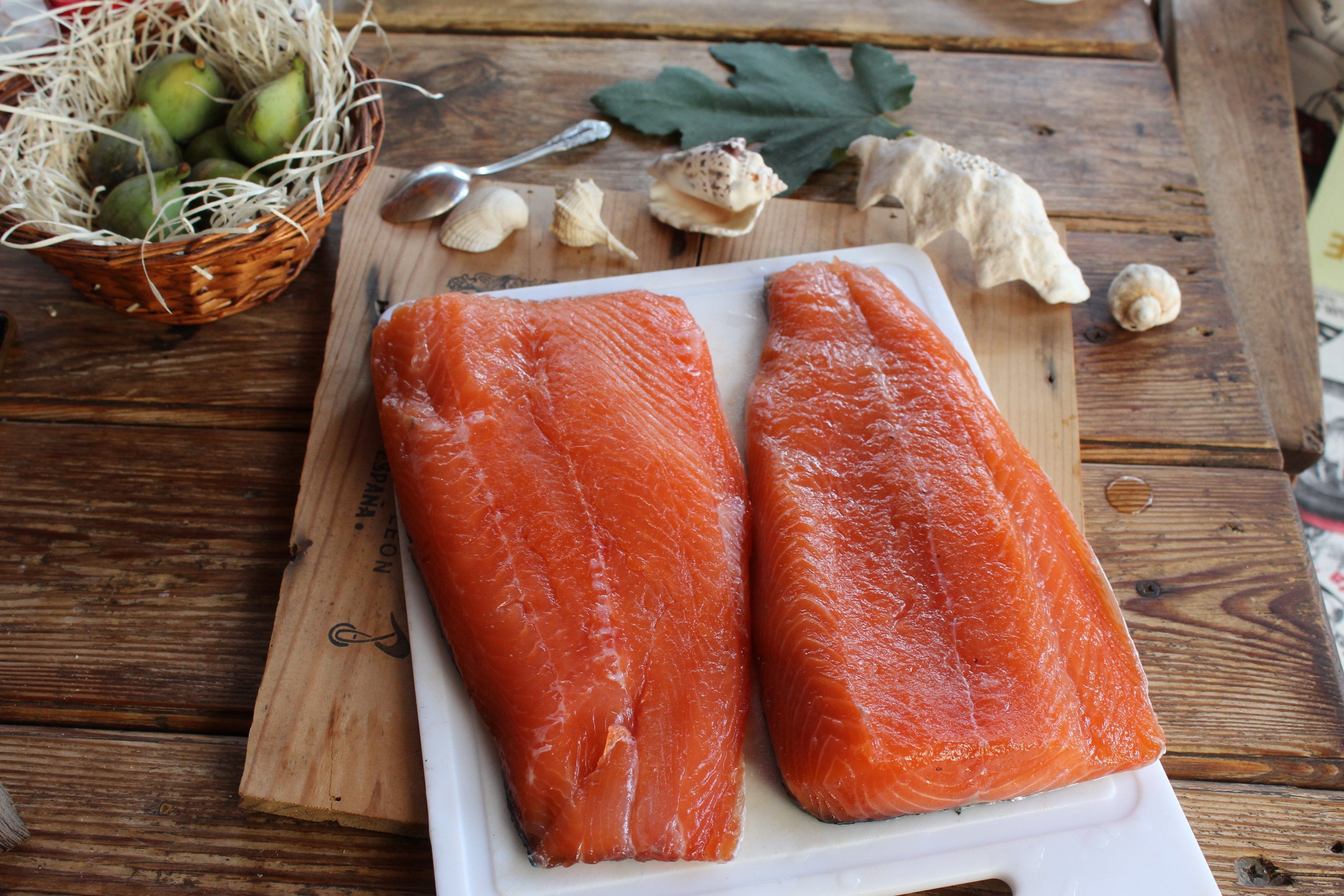 Filetes de salmón crudo sobre tabla blanca, listos para curado y ahumado casero, acompañados de elementos naturales como frutas verdes, conchas y hojas decorativas.