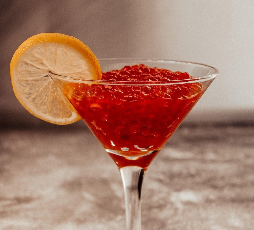 Copa de martini con falso caviar de sandía, decorada con rodaja de limón y acompañada de cuchara con esferas rojas.
