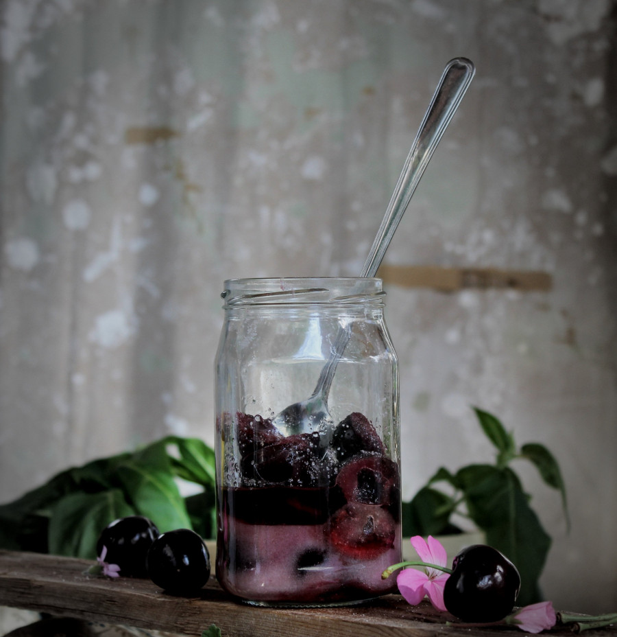 Tarro de licor Kirsch casero con cerezas frescas y azúcar, sobre mesa rústica con flores y utensilios de cocina