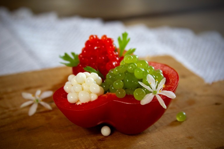 Mitades de pimiento rojo rellenas con esferas de caviar molecular en colores rojo, blanco y verde, decoradas con perejil y flores blancas sobre fondo rústico.