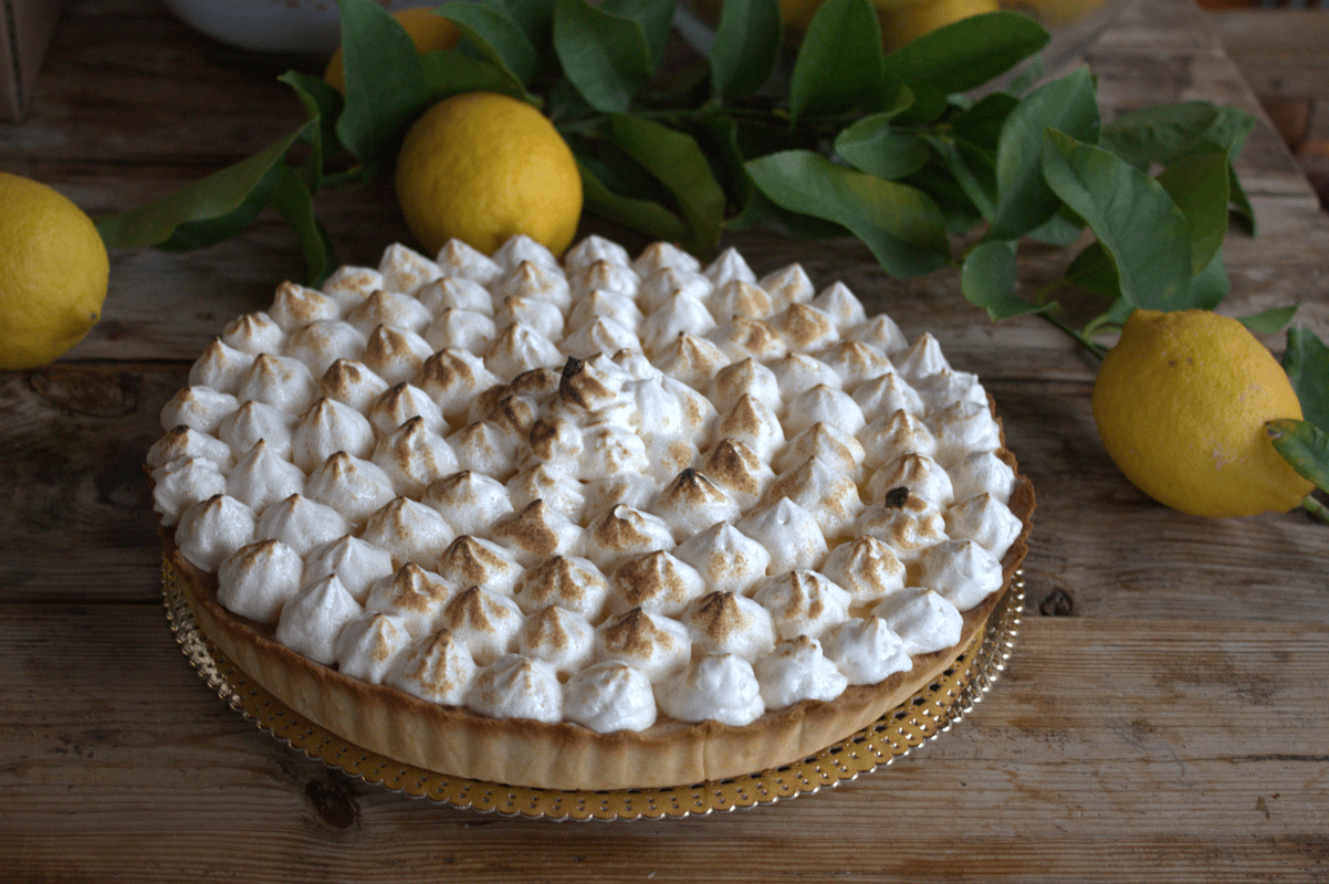 Lemon Cake (Tarta de Limón) 2 Tarta de limón con picos de merengue flambeados sobre una base de masa quebrada, rodeada de limones frescos con hojas en una mesa de madera.