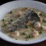 Merluza en salsa verde con gambas servida en un plato blanco con guisantes y ajos fritos.