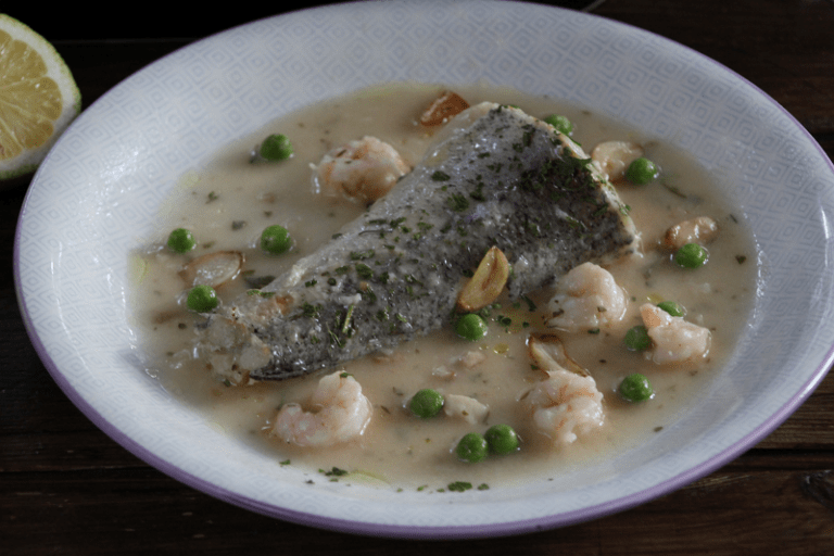 Merluza en salsa verde con gambas servida en un plato blanco con guisantes y ajos fritos.