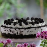 Cheesecake de Oreo sin horno completa sobre soporte rosa, decorada con nata y mini galletas, rodeada de geranios rosas desenfocados.