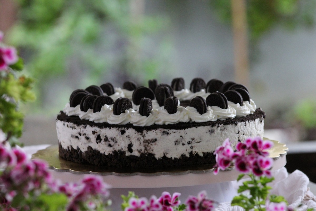 Cheesecake de Oreo sin horno completa sobre soporte rosa, decorada con nata y mini galletas, rodeada de geranios rosas desenfocados.